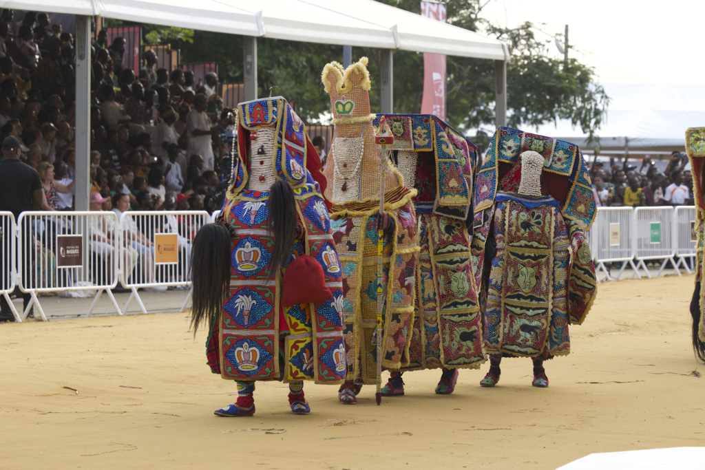 Four Egungun masquerades in beaded panels crossing the arena in front of the grandstand at the Festival des Masques