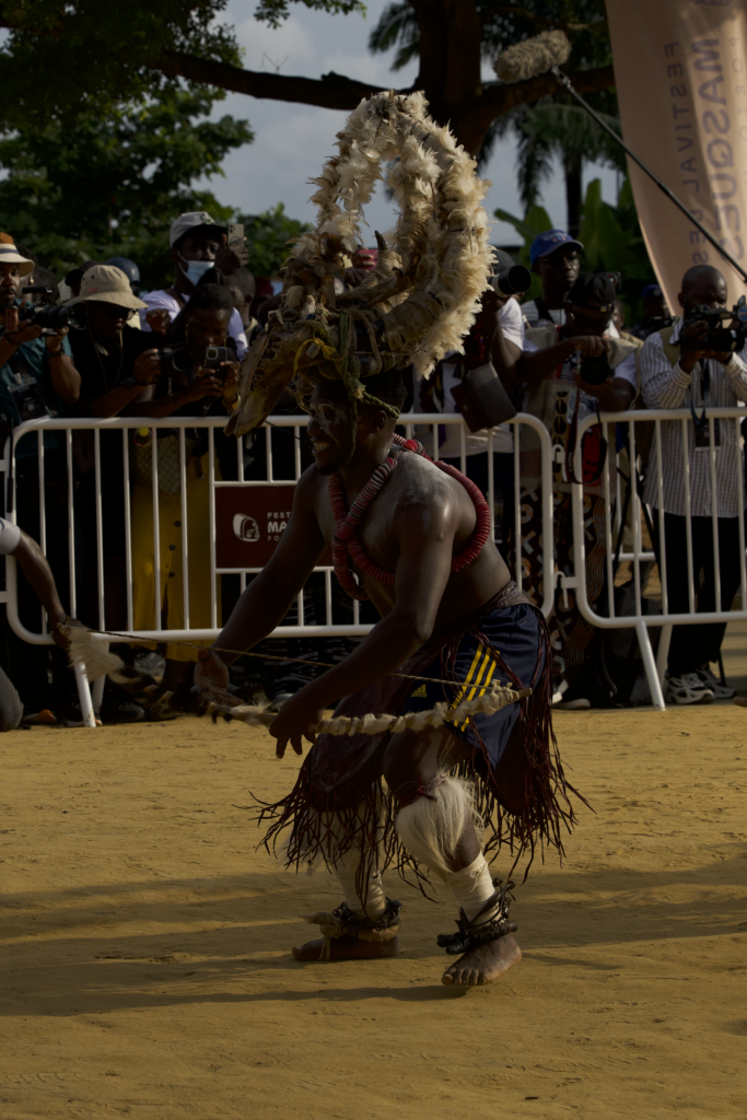 A masquerade dancer in a horned headdress performing in front of a barrier line of photographers