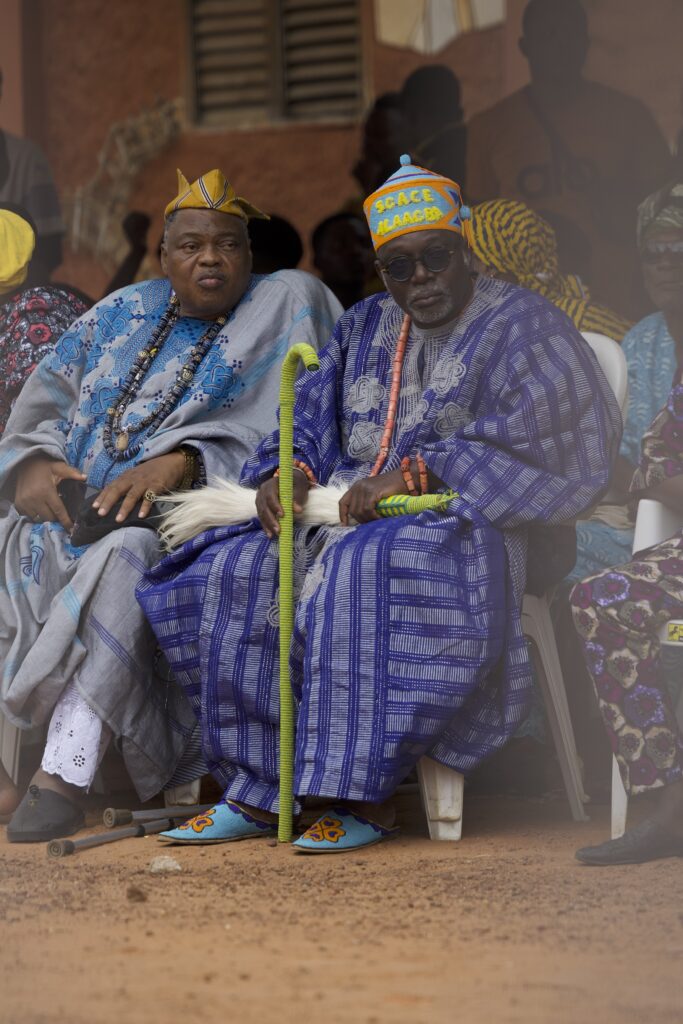 Elders in coral beadwork and indigo cloth seated as they wait for the masquerade procession, their regalia signalling rank