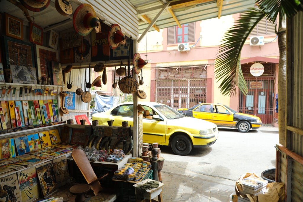 City Codes: Dakar Is Not a Trend 3 An art and crafts stall in Dakar hung with paintings, masks and drums, yellow taxis passing on the street outside.