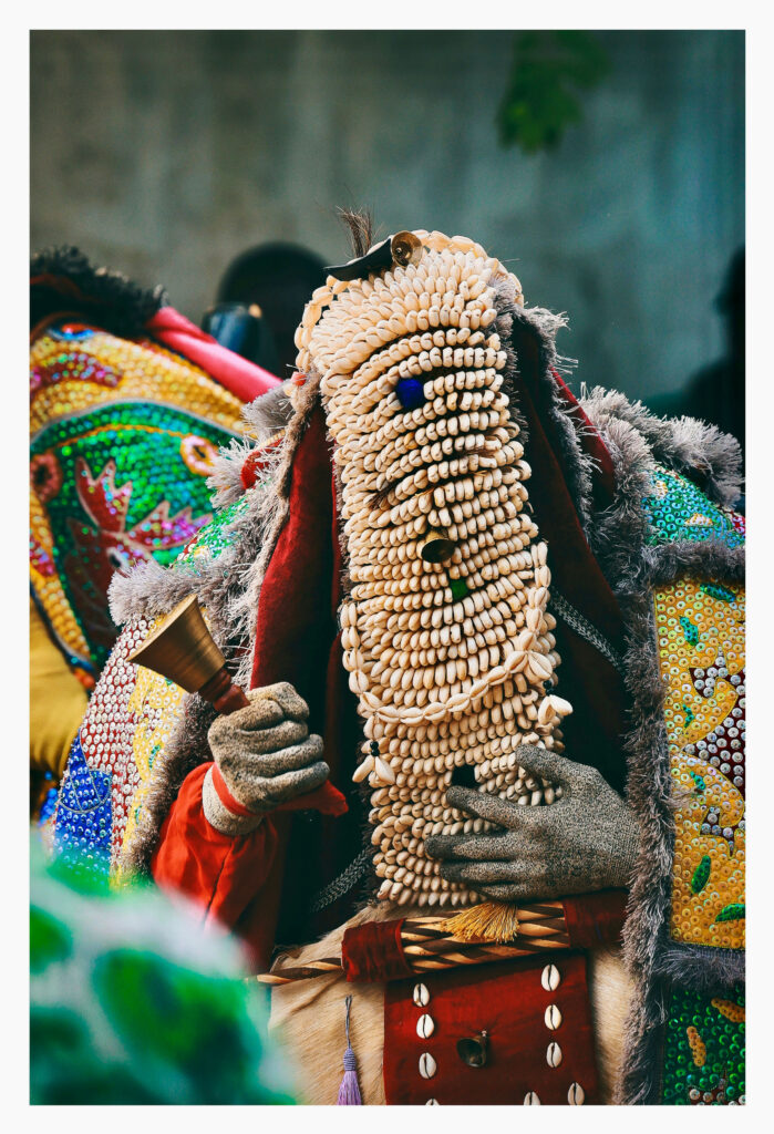 Masquerade costume covered entirely in cowrie shells, holding a bell in a gloved hand