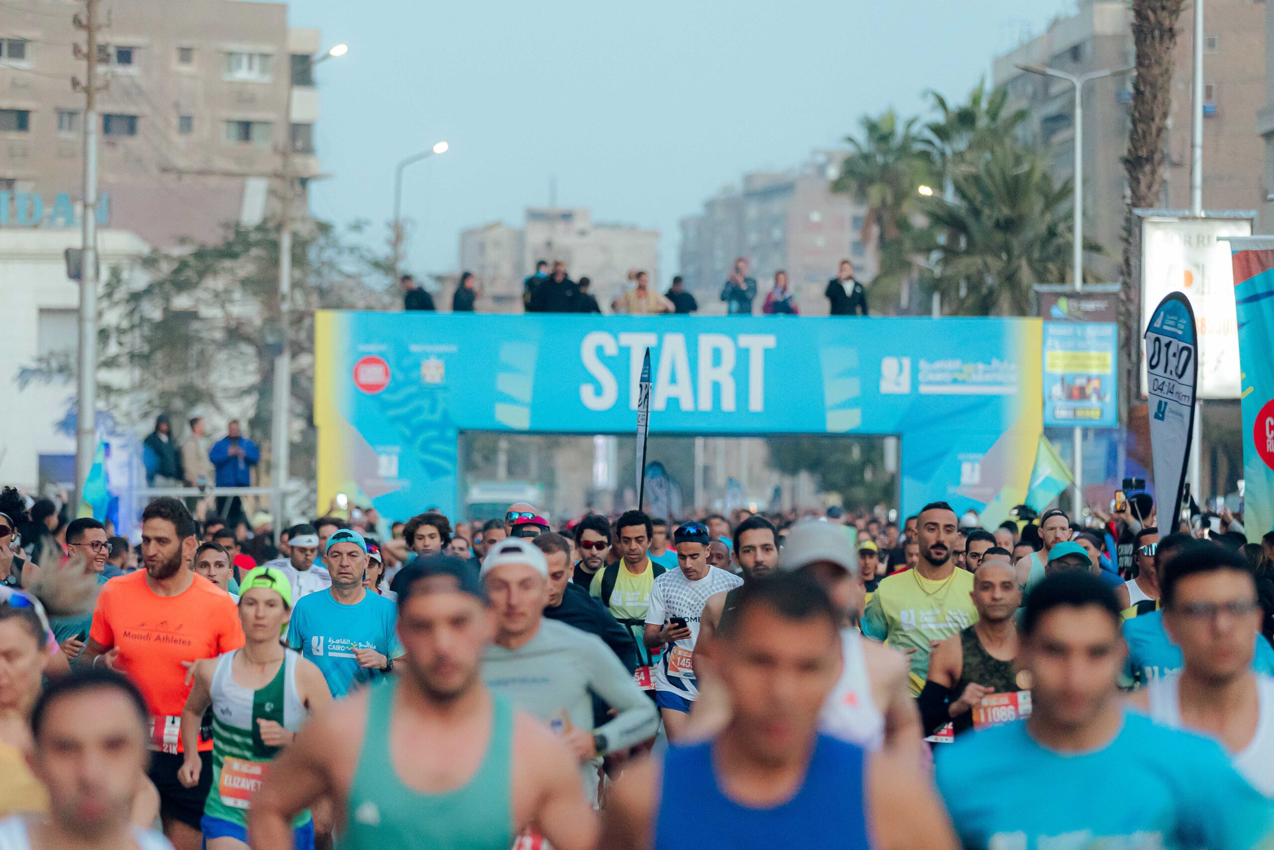 Runners packed beneath a blue START gantry at a city race, apartment blocks rising behind.