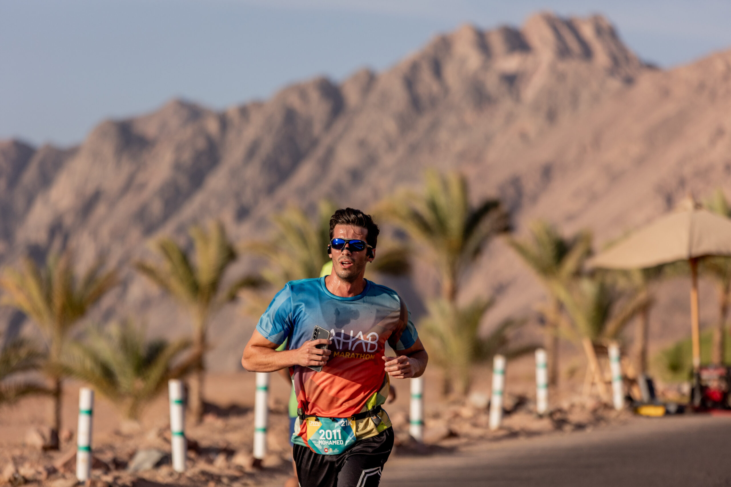 A runner in a blue race vest and sunglasses on a desert road, a bare rocky mountain and palm trees behind.