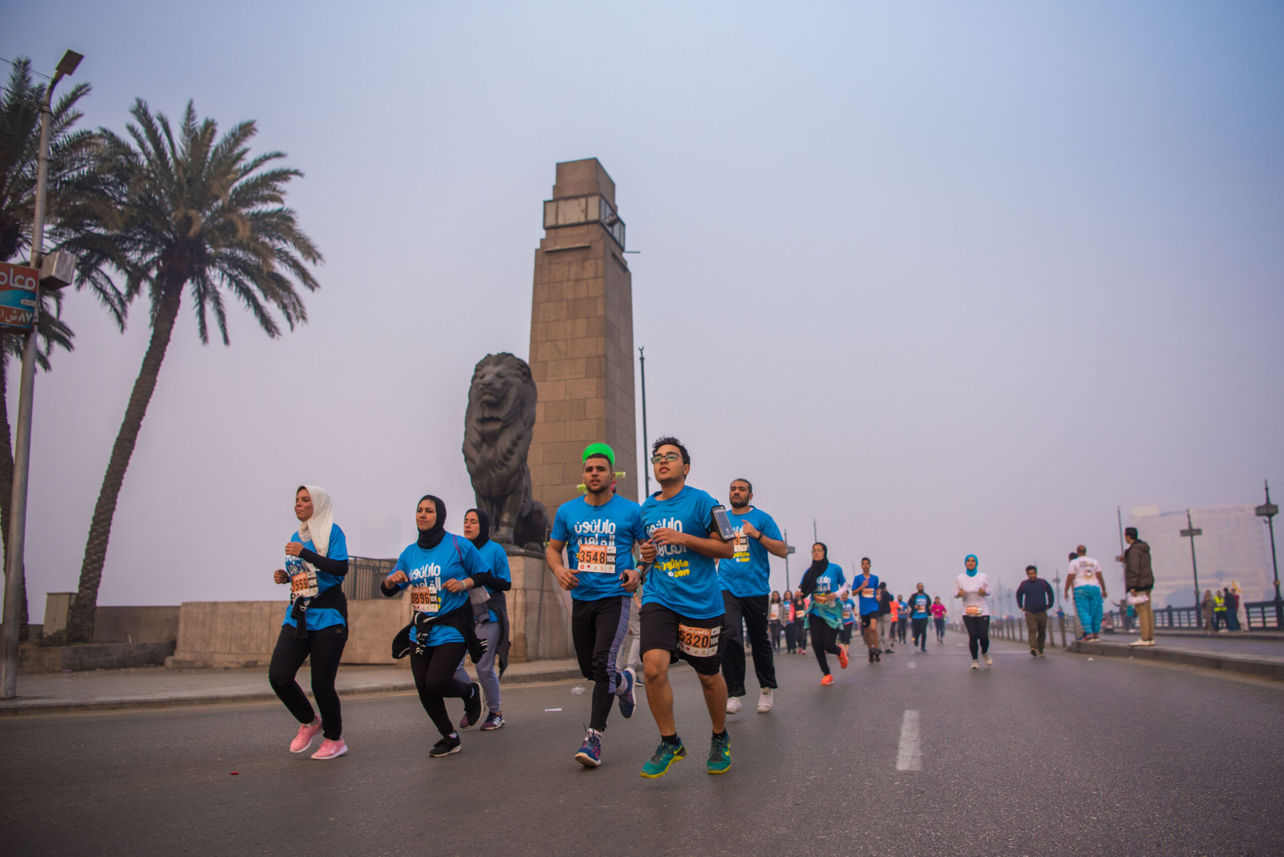 Runners on a wide road at dawn beside a tall stone monument and palm trees in hazy light.