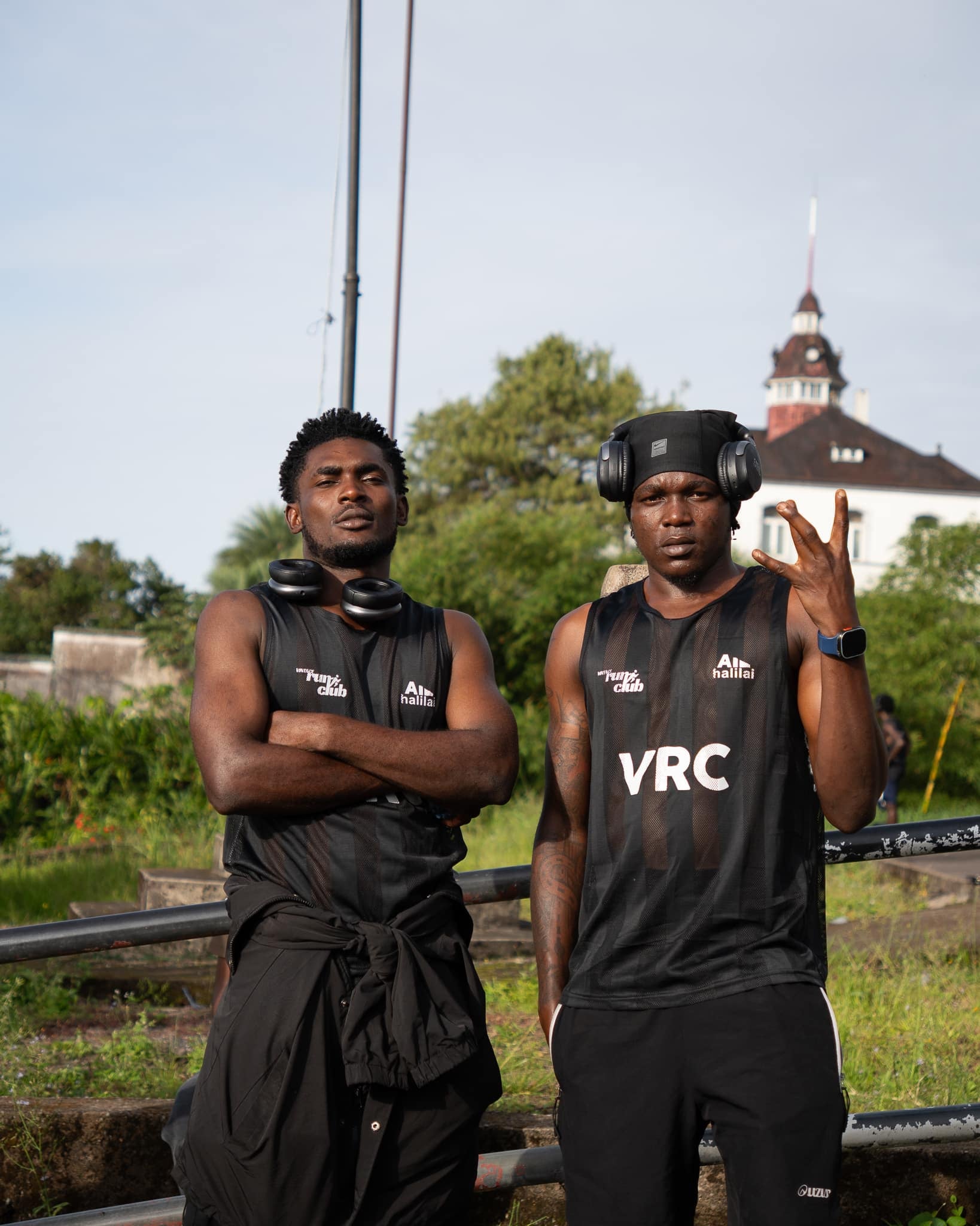 Two runners in black Vintage Run Club vests stand on a low wall, one with arms crossed, a green hillside and a red-roofed building behind.