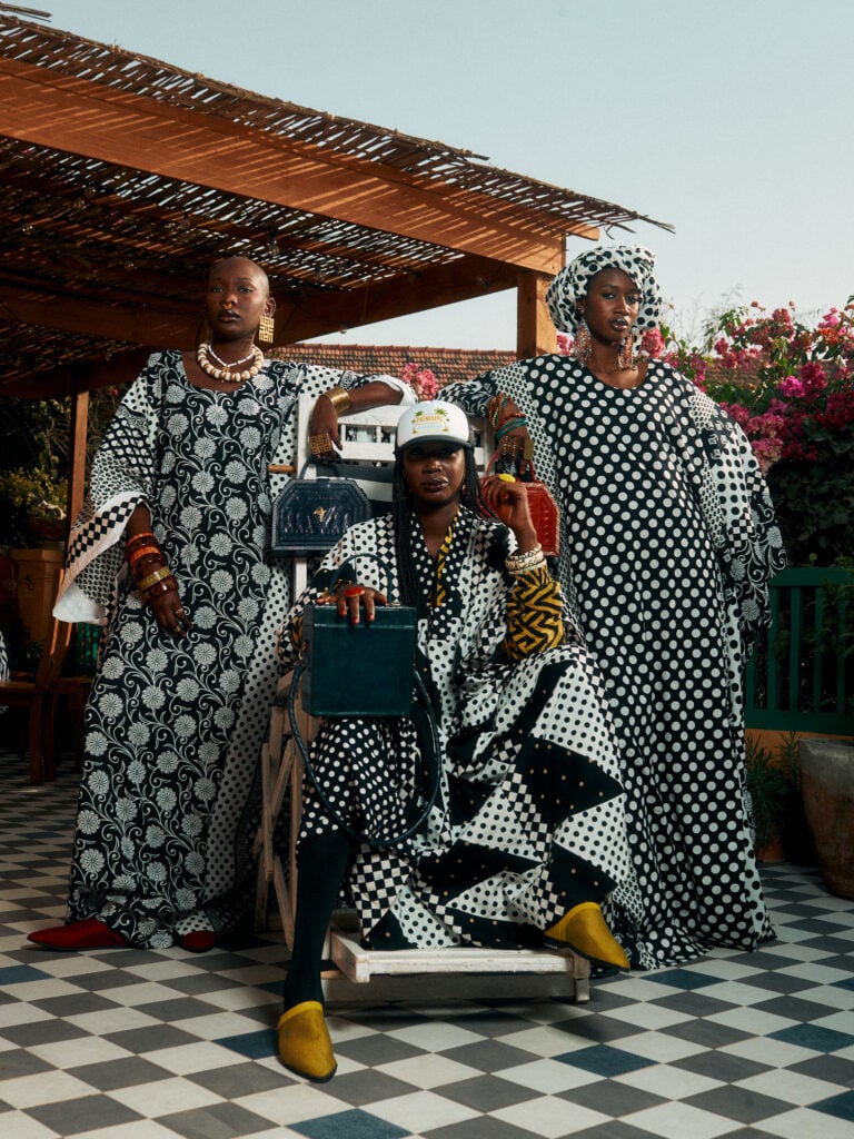 City Codes: Dakar Is Not a Trend 5 Three models in monochrome patterned boubous on a bougainvillea-lined rooftop terrace in Dakar, two standing and one seated in a white cap with structured handbags.