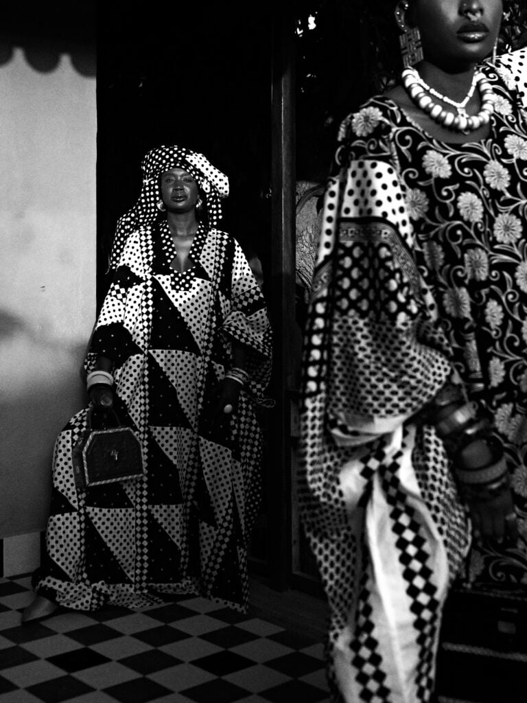City Codes: Dakar Is Not a Trend 2 A black-and-white portrait of two models in patterned boubous, one standing in a doorway with a box bag, the other close in a beaded necklace and bangles.