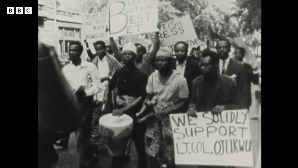 Marchers with a drum and hand-lettered placards, one reading We Solidly Support Lt. Col. Ojukwu, another addressing the British press.