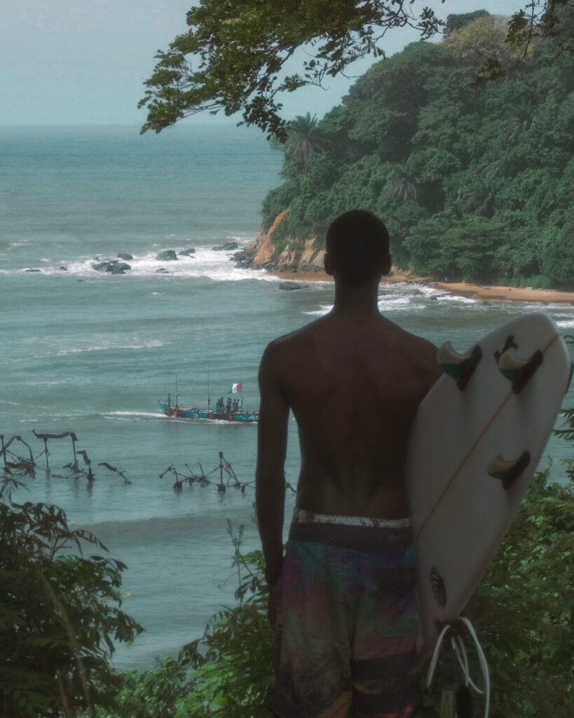 A surfer holding his board looks down over a forested cove in Côte d’Ivoire as a fishing pirogue passes below