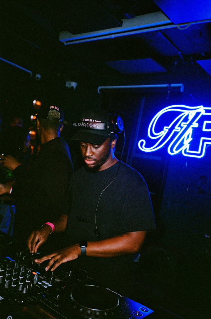 How DJs Helped Take Afrobeats Global 3 DJ Selecta Suave working the mixer in a black cap and T-shirt beside a blue neon sign
