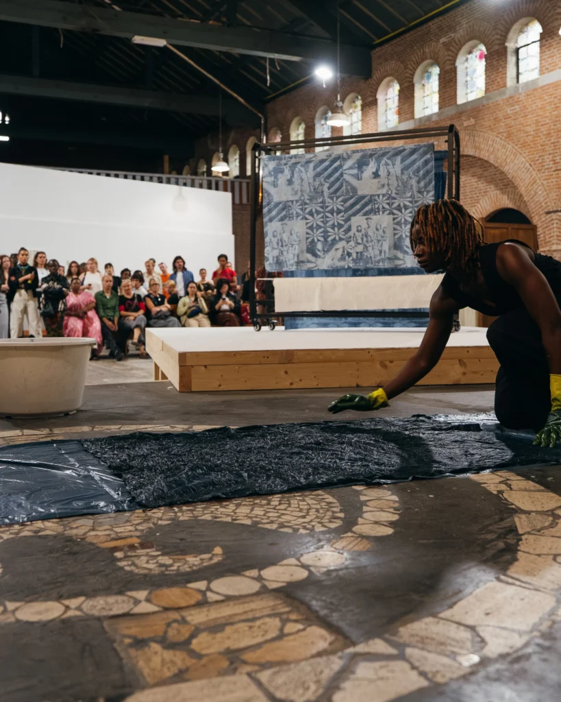 Adirẹ and the Wrapper-Wearers 2 Kosisochukwu Nnebe kneels in yellow gloves to spread a large indigo cloth across a stone floor as an audience watches from tiered seating.