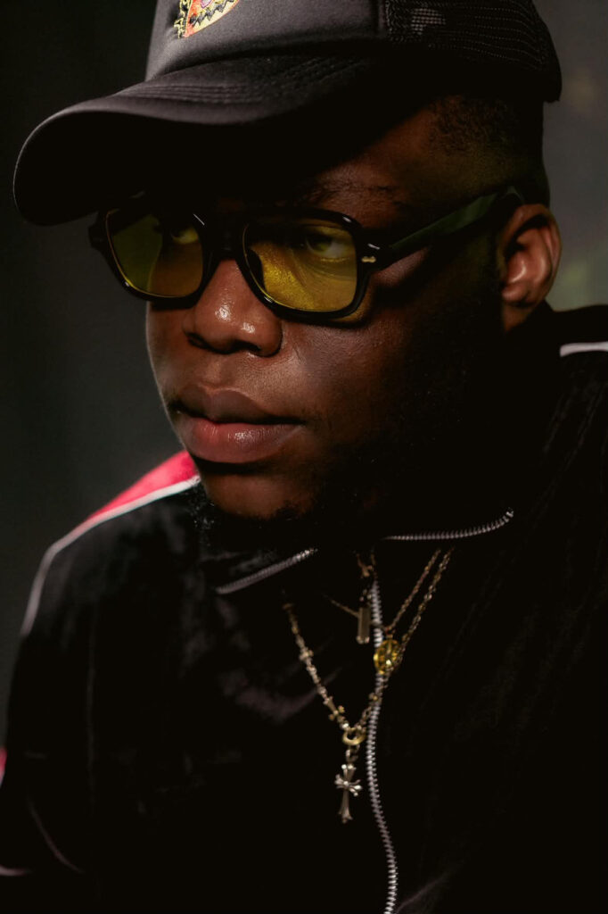 How DJs Helped Take Afrobeats Global 1 Close portrait of DJ Trill Xoe in a black cap, yellow tinted sunglasses and layered gold chains