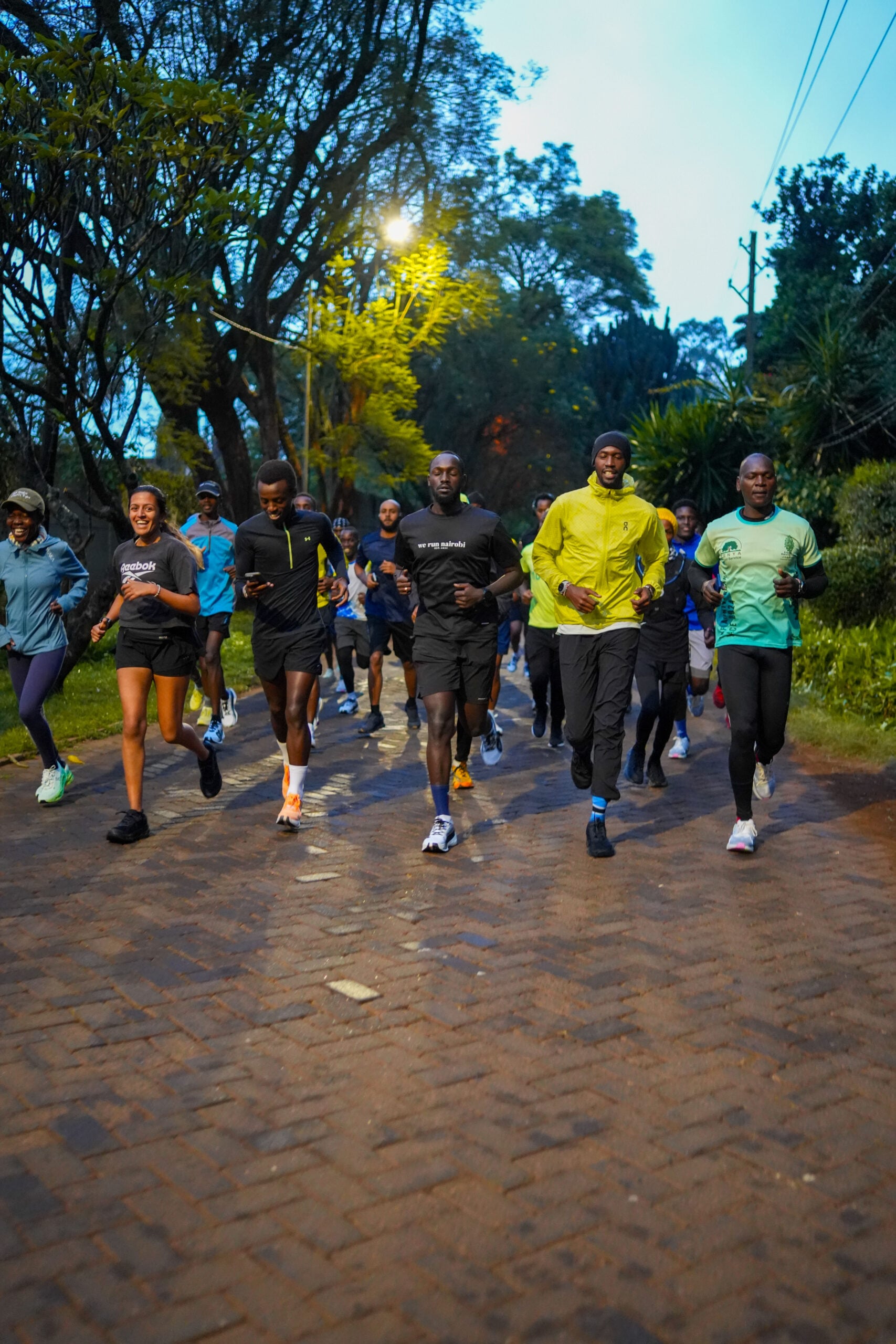 A group of runners on a tree-lined tarmac road at first light, moving together toward the camera.
