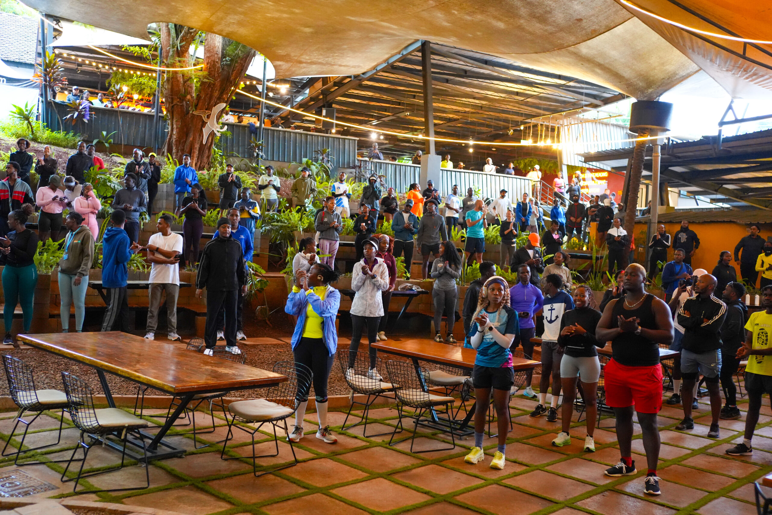 Long wooden communal tables in the foreground of a covered courtyard, a crowd of runners standing and talking behind them.