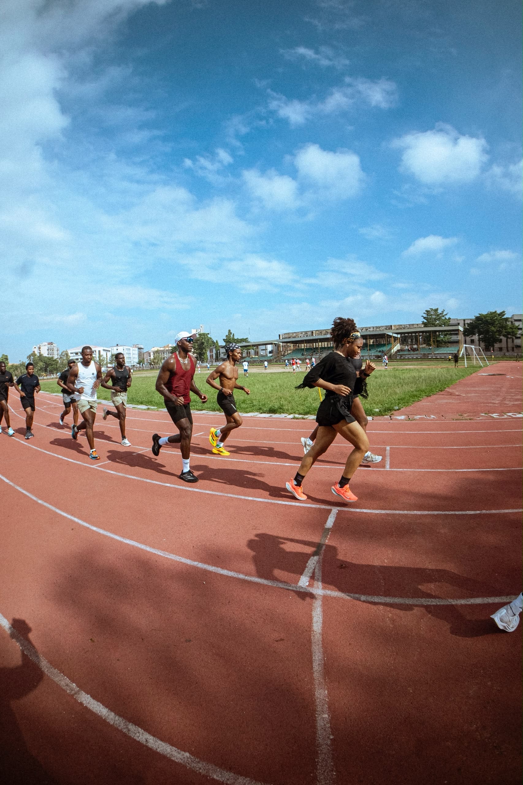 Runners on a red athletics track under a bright blue sky, a woman in black leading in the foreground.