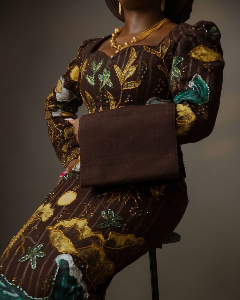 Can a Piece of Clothing Bring You Home? 3 Cropped studio portrait of a seated woman in a fitted brown gown of embroidered cloth with gold and green floral appliqué, a folded George wrapper resting across her lap.