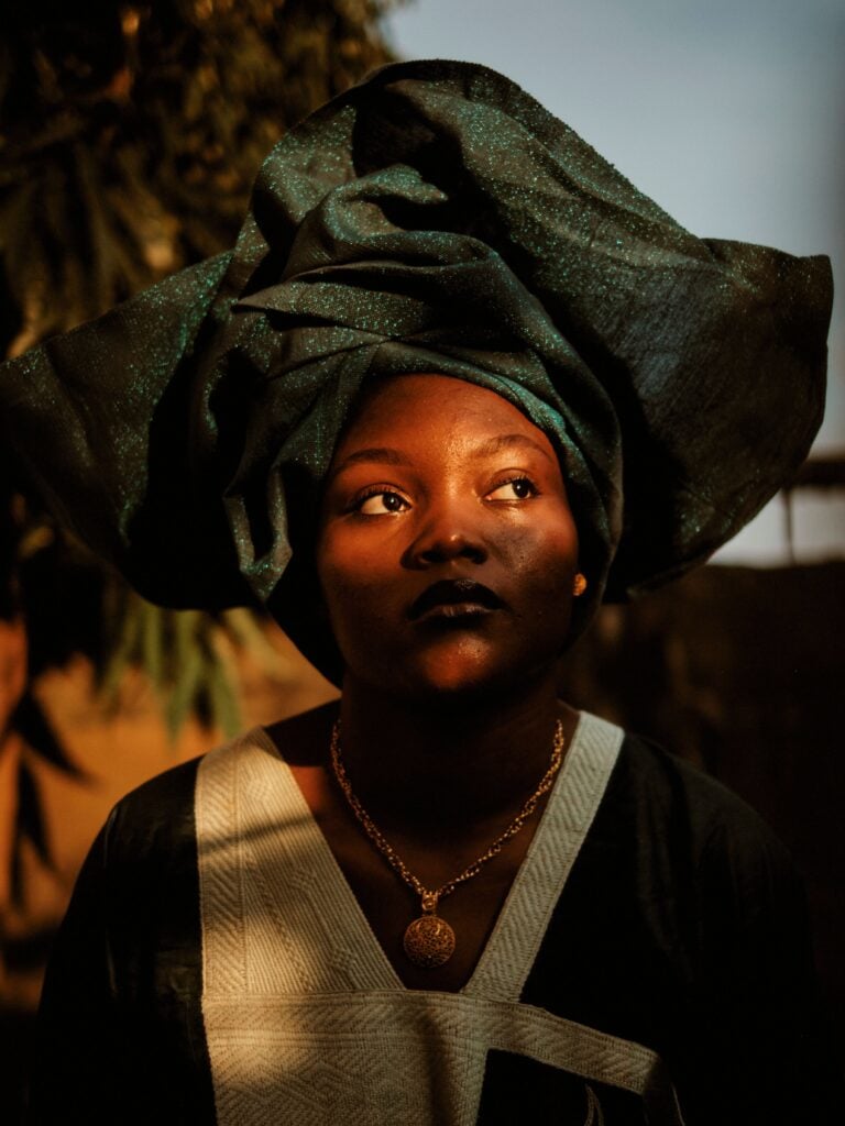 Can a Piece of Clothing Bring You Home? 2 A woman in a large sculptural gele of dark teal shimmering cloth, tied high and wide, worn over a dark tunic trimmed with cream.