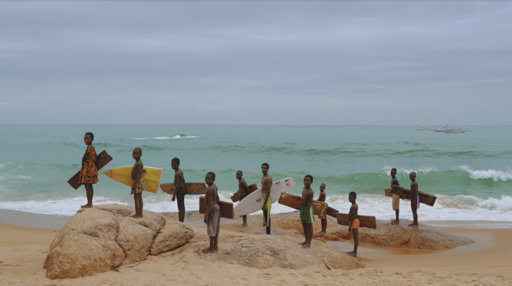 A line of young surfers stands on beach rocks in Côte d’Ivoire holding surfboards and carved wooden boards