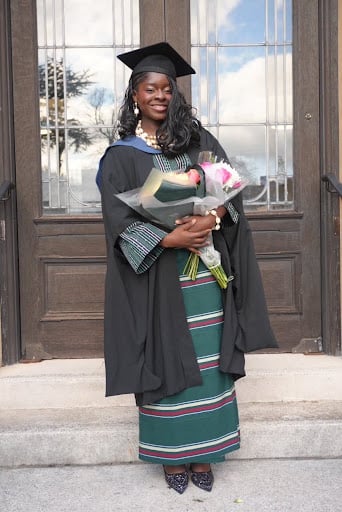 Can a Piece of Clothing Bring You Home? 1 Tola stands in front of a panelled door in a black graduation gown and mortarboard worn over a floor-length green, white and pink handwoven Aṣọ òkè wrapper, holding a bouquet of flowers.