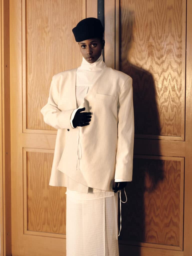 Portrait in a cream tailored suit and black gloves against a wood-panelled door