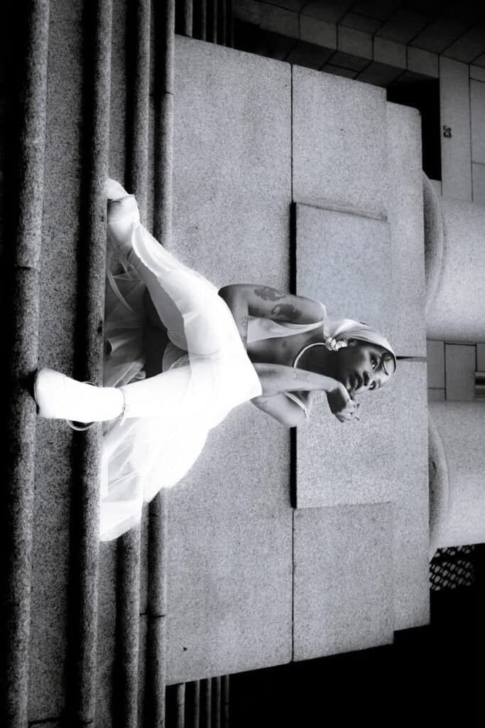 Figure dressed in white posed against brutalist concrete blocks