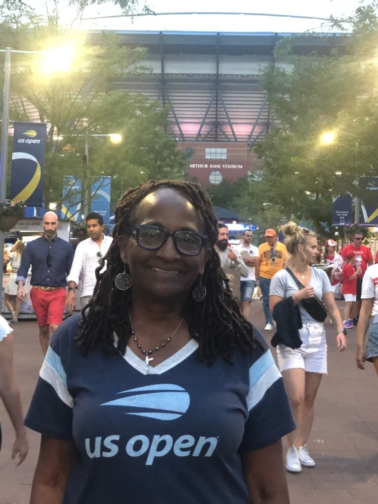 The author's mother smiling in a US Open t-shirt at Flushing Meadows, with the floodlit Arthur Ashe Stadium and its name sign behind her.