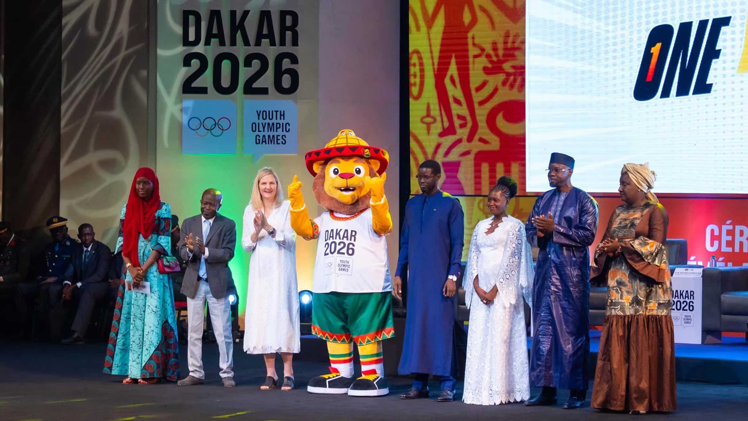 Who Made the Welcome 4 The Ayo mascot of the Dakar 2026 Youth Olympic Games on stage at its unveiling ceremony in Senegal, flanked by officials