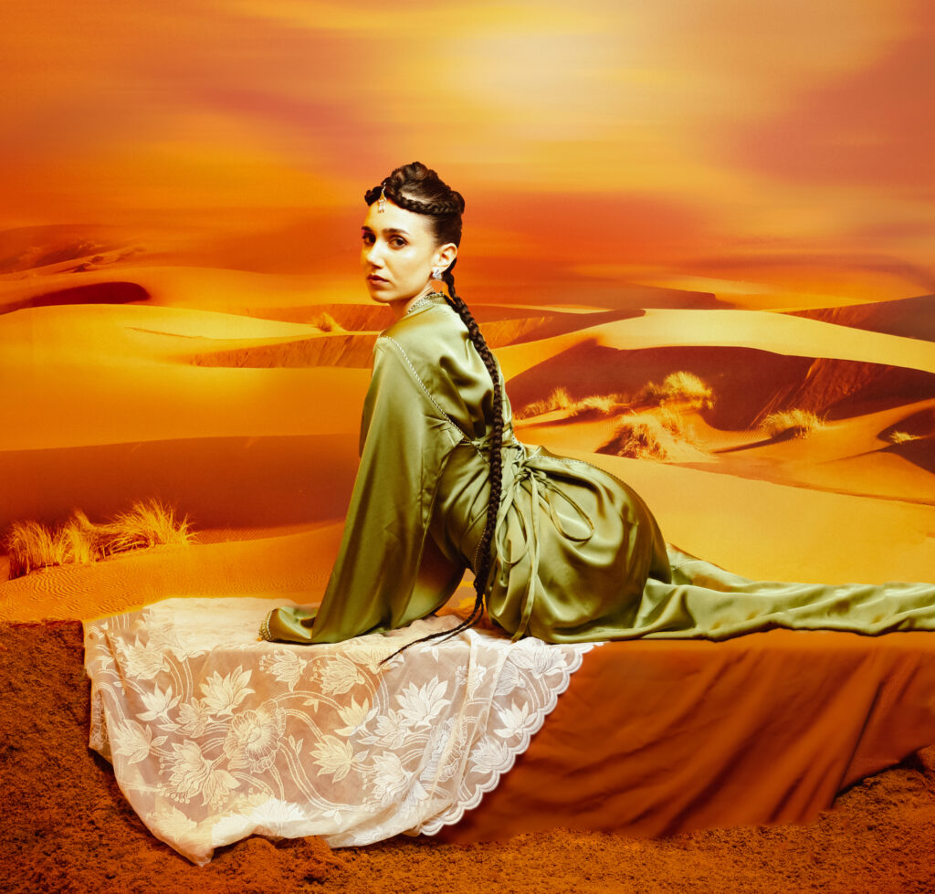 North African Women Wore the Future First 5 A model in a green YSR Maison velvet gown seated on lace in an orange desert, styled with Tausa World jewellery.