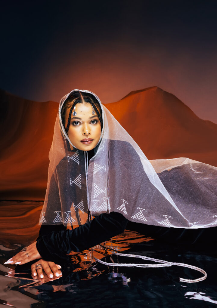 North African Women Wore the Future First 2 A model kneeling in a sheer patterned veil over a black Ruaa Elmansuri look, hands in shallow water in a dusk desert.