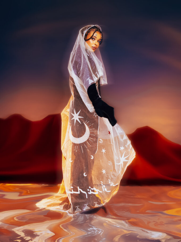 North African Women Wore the Future First 1 A model standing draped in a sheer veil printed with stars and crescent moons over a black Ruaa Elmansuri look, in a dusk desert with water underfoot.