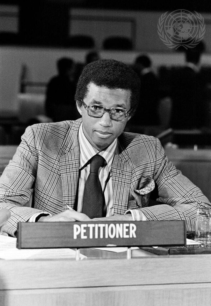 Arthur Ashe seated behind a PETITIONER nameplate as he addresses the United Nations Special Committee on Apartheid in 1970, in black and white.