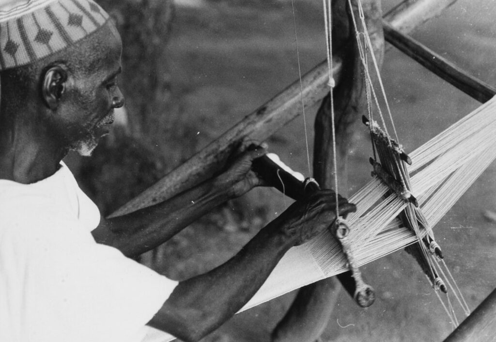 From Korhogo Cloth to Digital Print: Alia Baré and the Language of Senufo Textiles 1 Black-and-white photograph of a man in a patterned cap working a narrow-strip handloom outdoors near Korhogo, 1966.