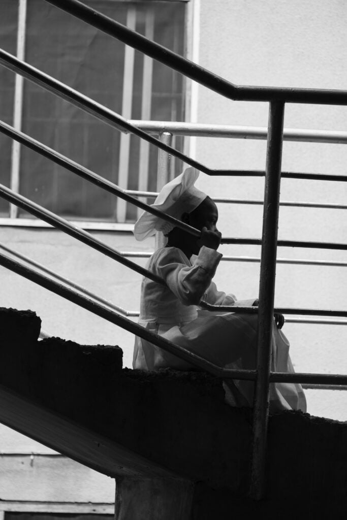 Holy Dress Codes: What Comes to Mind When You See the Garment? 8 A worshipper in a white Aladura garment sits alone on a concrete stairway behind metal railings, in black and white by Stephen Tayo.