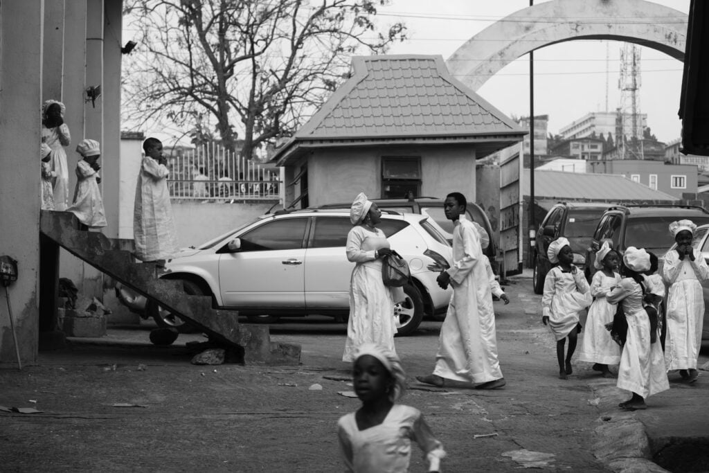 Holy Dress Codes: What Comes to Mind When You See the Garment? 1 A worshipper in a white Aladura garment sits alone on a concrete stairway behind metal railings, in black and white by Stephen Tayo.