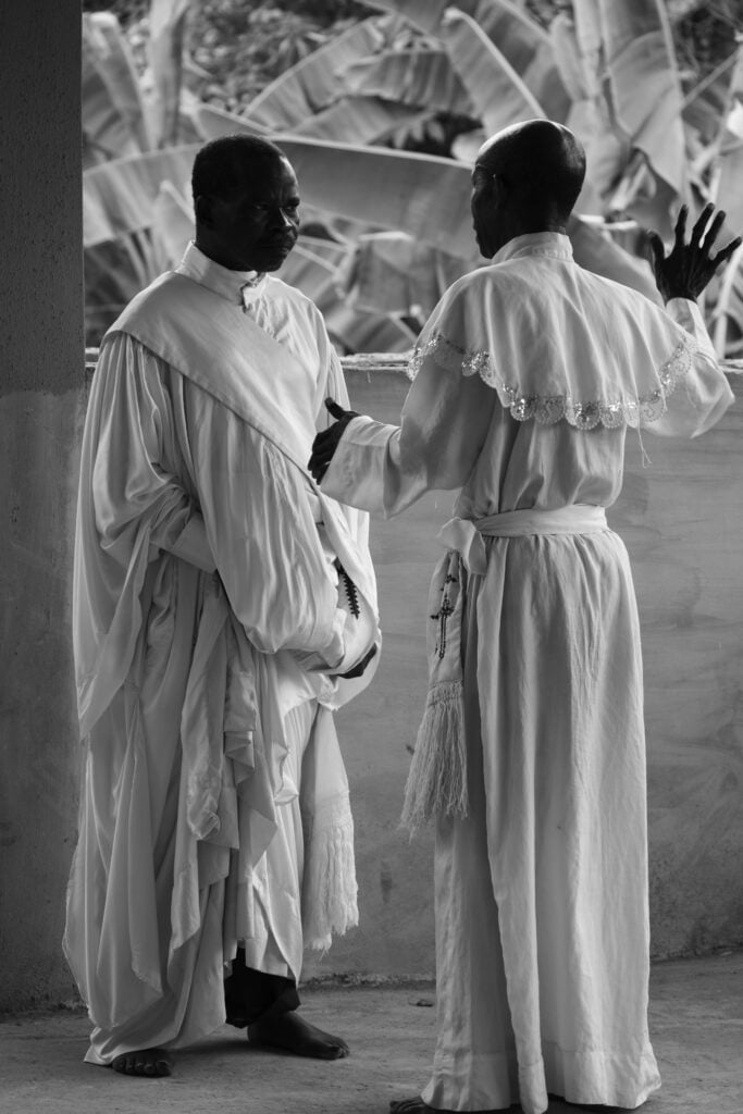 Holy Dress Codes: What Comes to Mind When You See the Garment? 2 Two men in white Aladura garments in close conversation, one wearing a tasselled cord, banana leaves behind them, in black and white by Stephen Tayo.