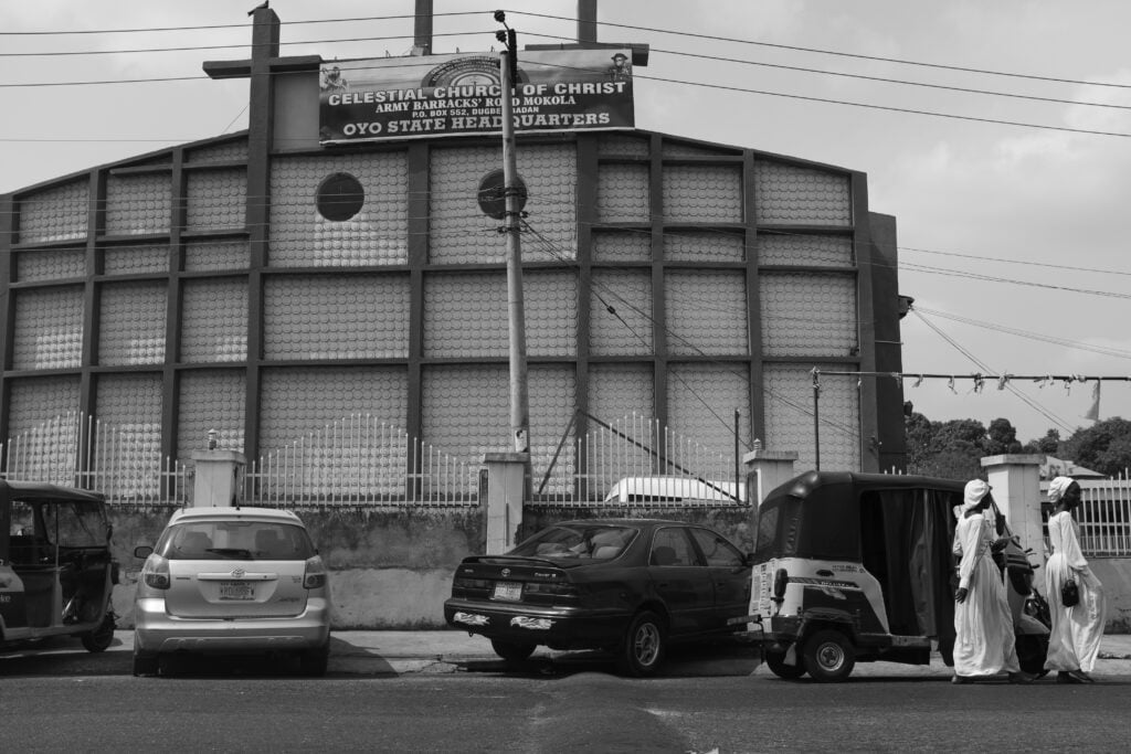Holy Dress Codes: What Comes to Mind When You See the Garment? 3 The Celestial Church of Christ headquarters in Mokola, Ibadan, with parked cars, a keke and figures in white garments on the street, in black and white by Stephen Tayo.
