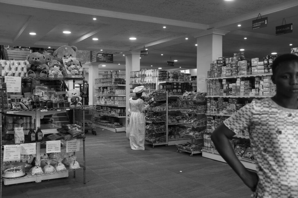Holy Dress Codes: What Comes to Mind When You See the Garment? 4 A woman in a white Aladura garment shops in a supermarket aisle as a young person looks toward the camera, in black and white by Stephen Tayo.