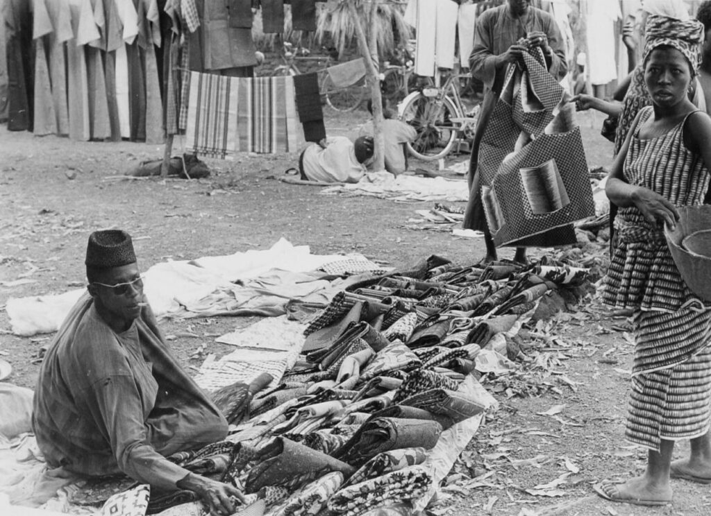 From Korhogo Cloth to Digital Print: Alia Baré and the Language of Senufo Textiles 3 Black-and-white photograph of a textile seller seated beside folded cloths laid out on the ground at Korhogo market, 1966.