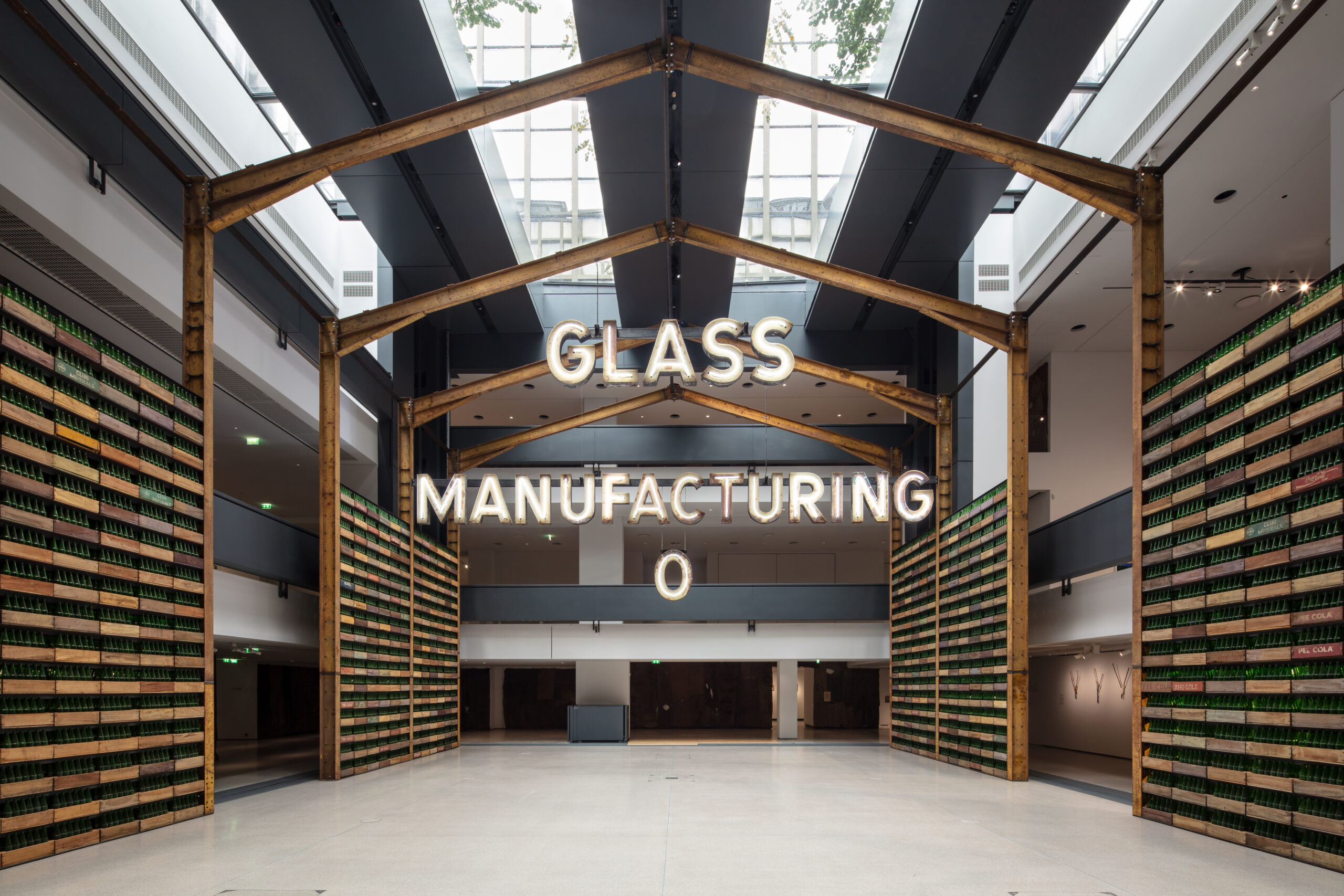 Ibrahim Mahama Brings Ghana's Industrial Past to Paris 1 A steel factory frame beneath an illuminated “GLASS MANUFACTURING” sign, flanked by shelves of green glass bottles.