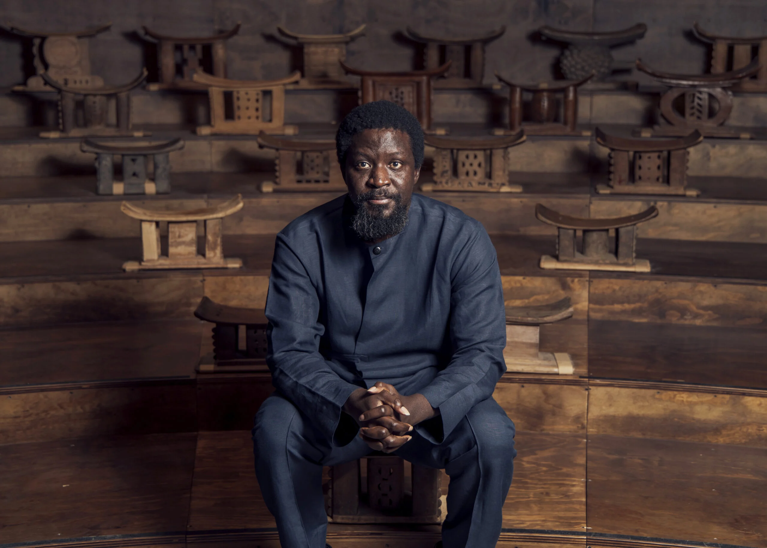 Ibrahim Mahama Brings Ghana's Industrial Past to Paris 4 Ibrahim Mahama dressed in dark blue, seated among wooden Asante stools arranged on stepped seating.
