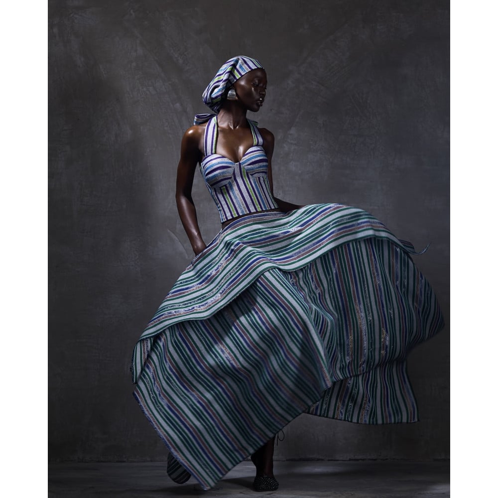 Model in a blue and green striped structured bodice and voluminous layered skirt with matching headwrap, photographed against a dark studio background.