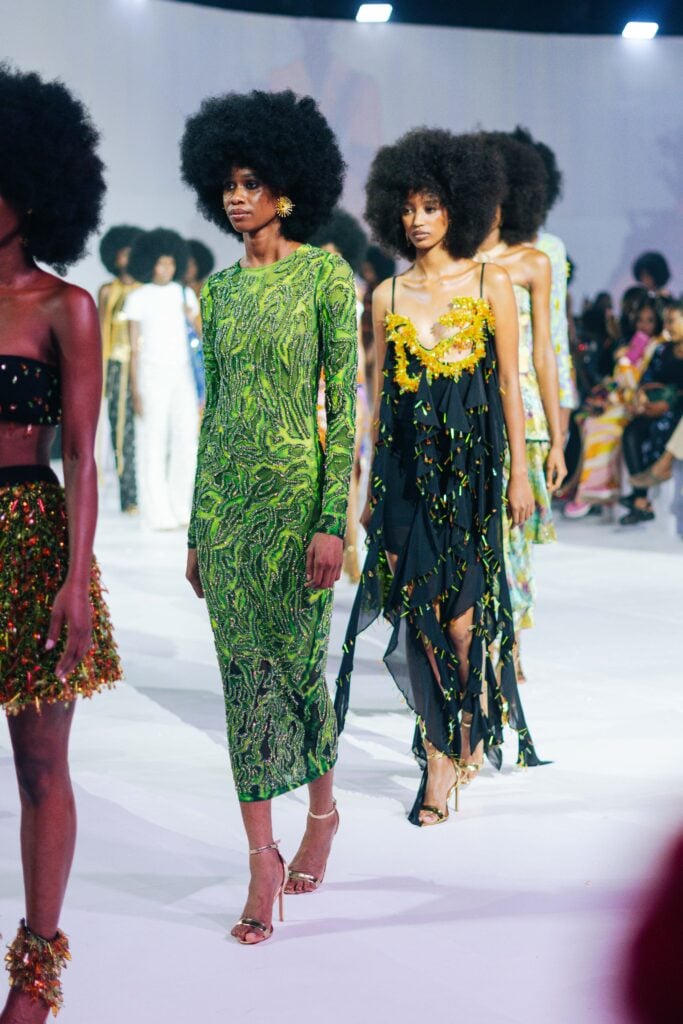 Models walk the runway in embellished Onalaja dresses, including a long-sleeved green dress and a black dress with yellow floral detailing.
