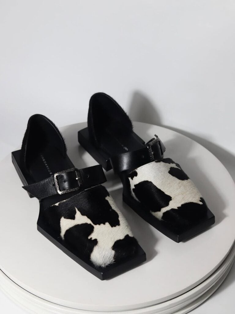 A pair of black, square-toe block heel shoes featuring a white cow-print pattern and a buckled strap, displayed on a white circular platform.