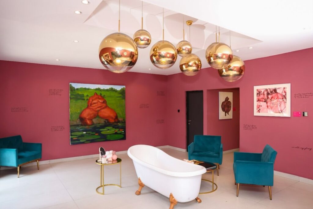 Uthman Wahaab’s paintings in a pink room with a white bathtub, teal chairs and gold pendant lights at Windsor Gallery, Lagos.