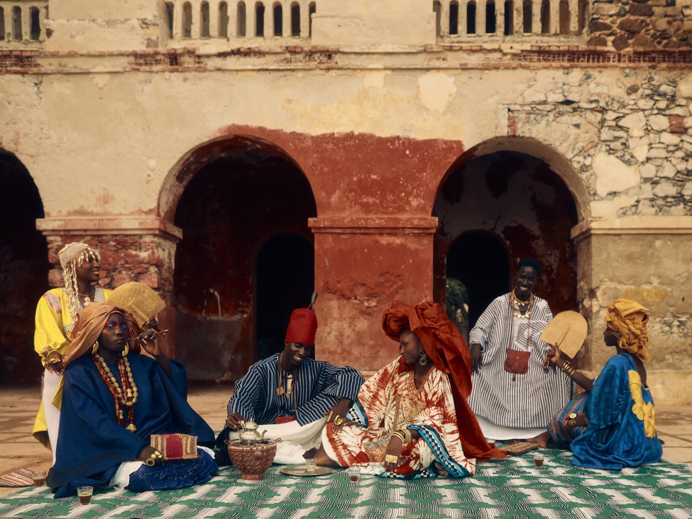 Six people in colourful clothing gather around a tea service in an arched courtyard for The Ritual of Attaya, the opening portfolio of Guzangs Atlas: Dakar.