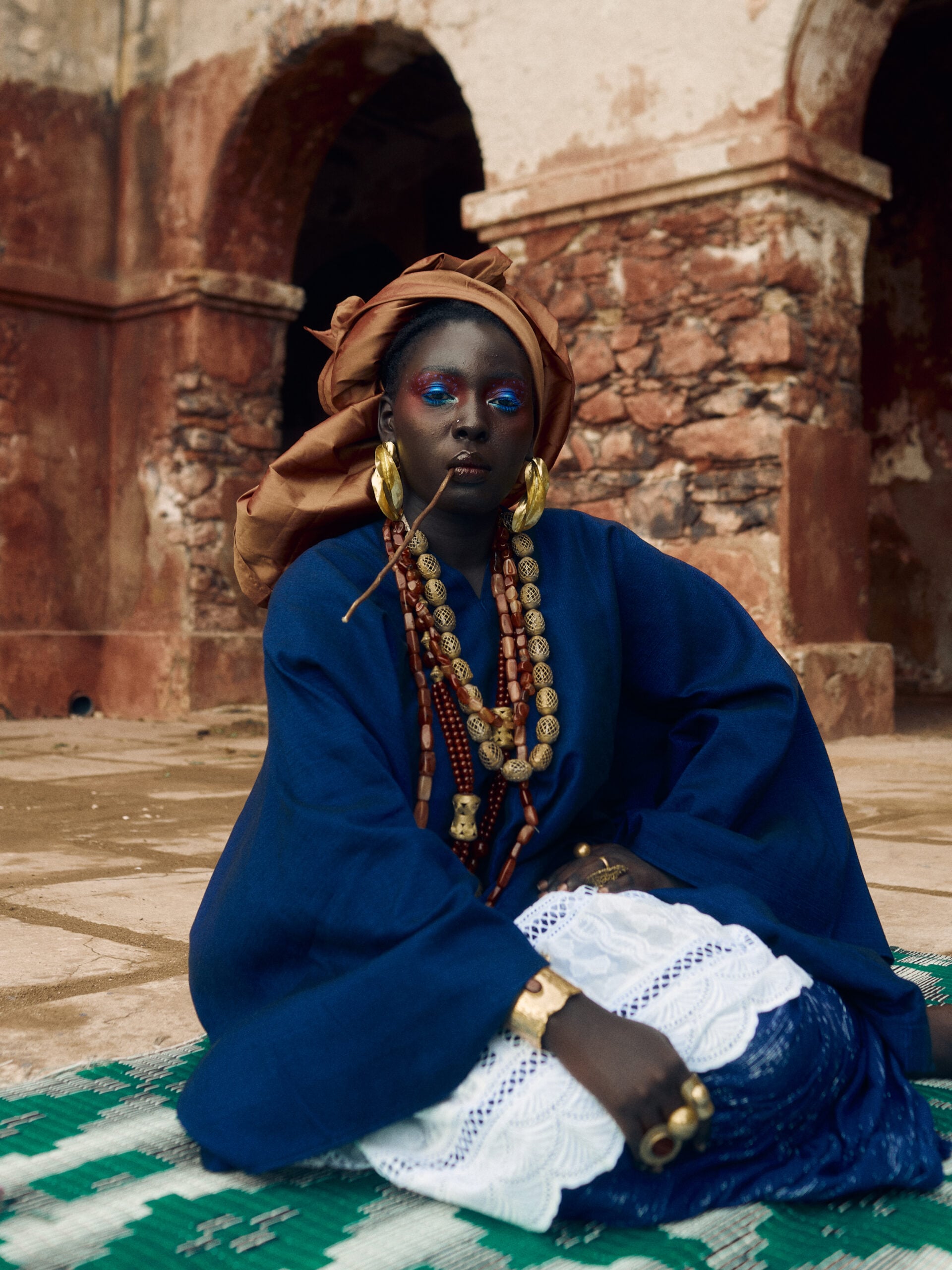 Woman in an indigo ensemble, rust-gold headwrap and layered necklaces seated beneath the terracotta courtyard arches on Gorée.
