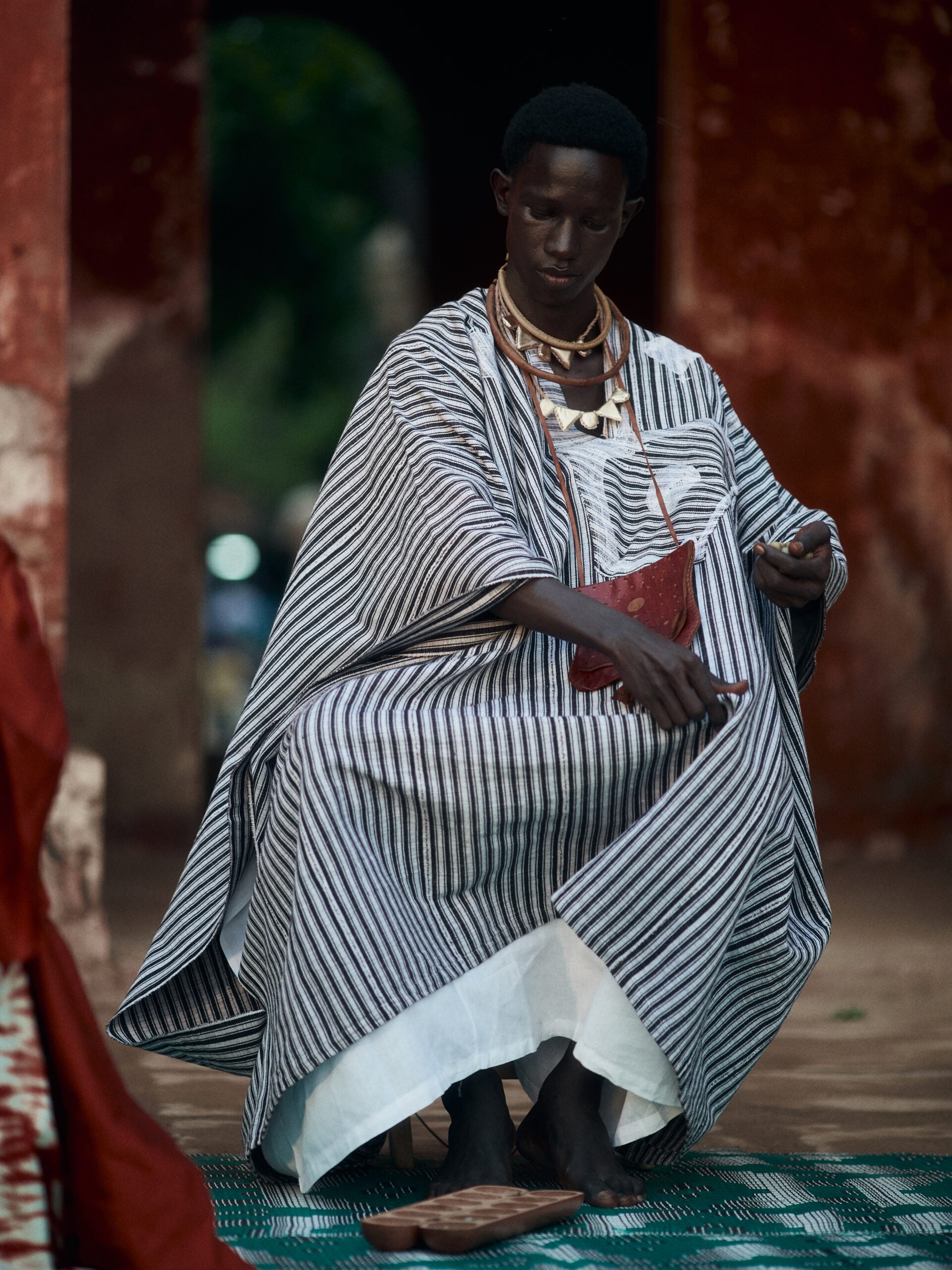 Man in a grey-striped mboubou, amulet necklaces and a red leather pouch sits beside an awalé board in the courtyard.