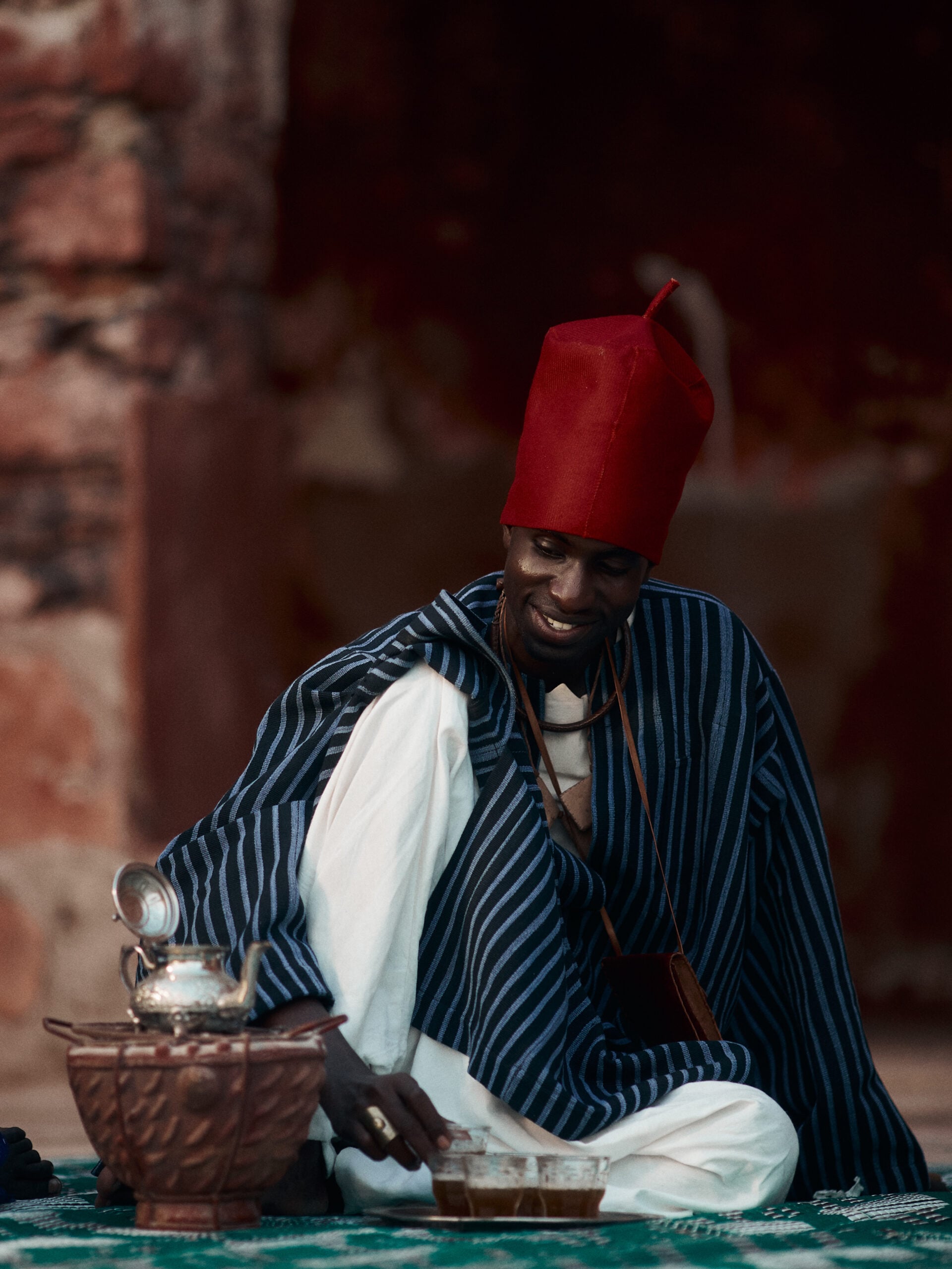 Tea host in a tall red cap and blue-striped robe seated beside a clay brazier, silver teapot and tray of attaya glasses.