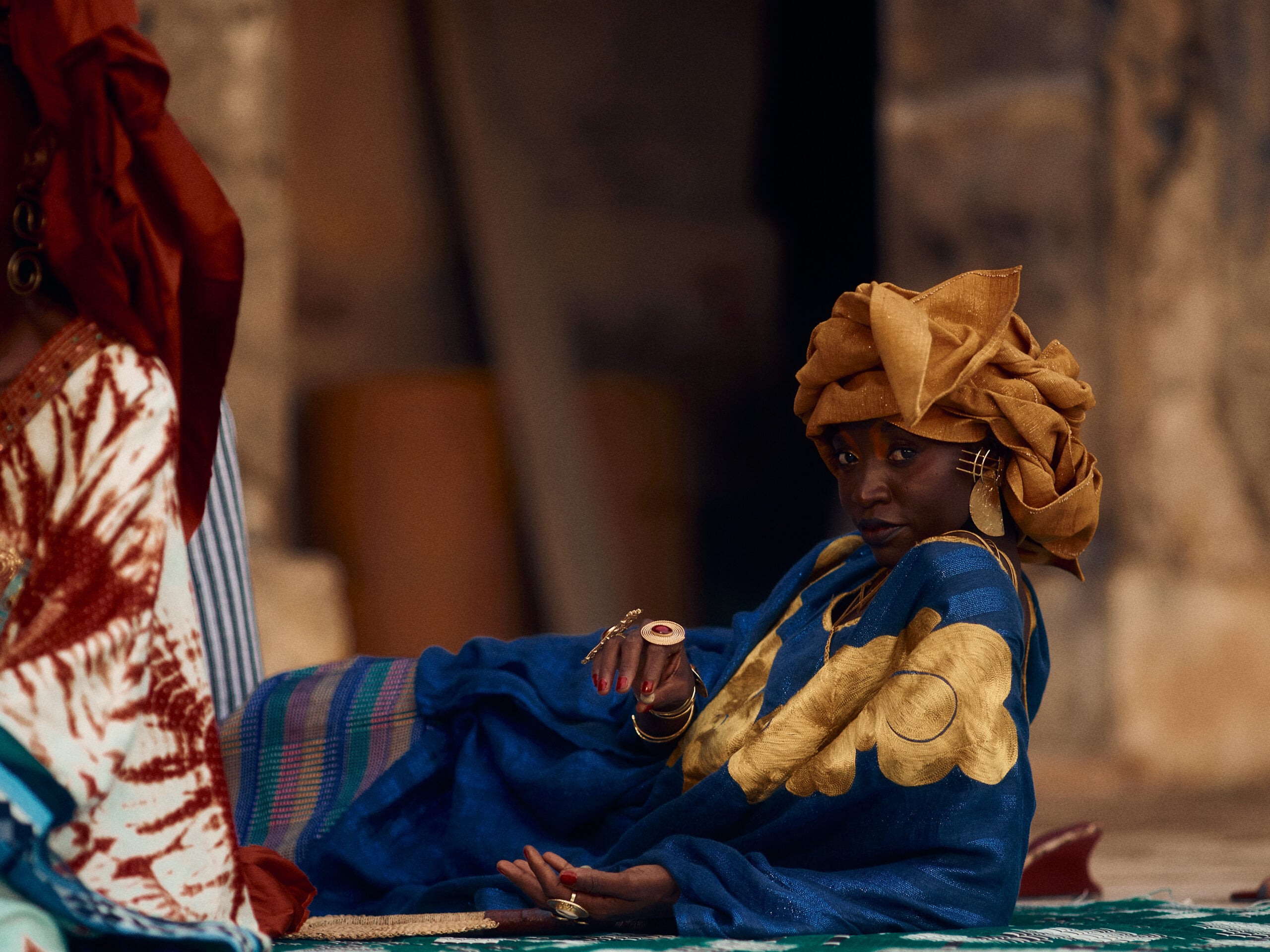Reclining woman in blue cloth with large gold floral motifs, a gold headwrap and sculptural jewellery on the woven basang.