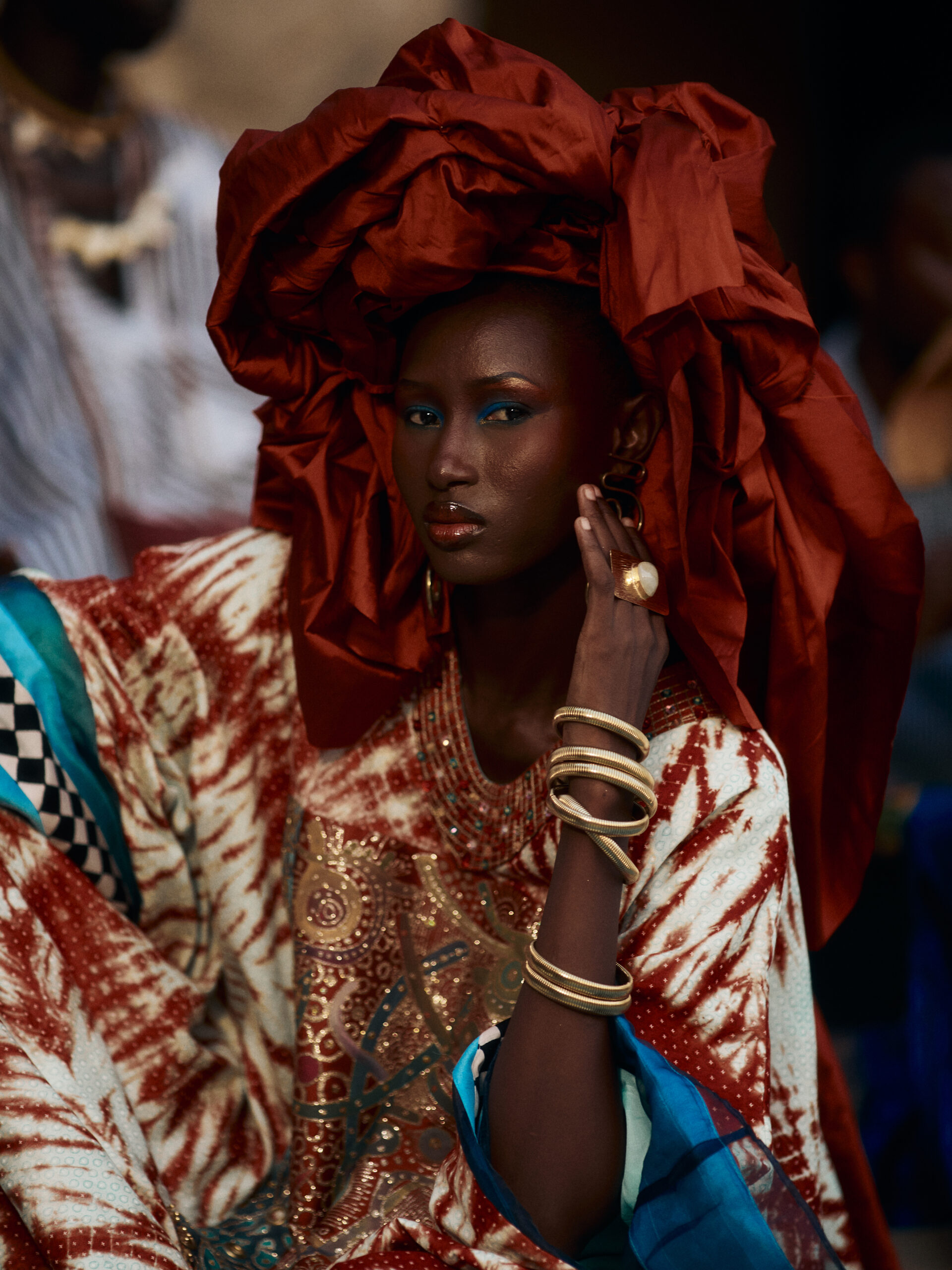Woman in a sculptural rust moussor and patterned mboubou, with her hand beside her face and gold-toned bracelets stacked along her arm.